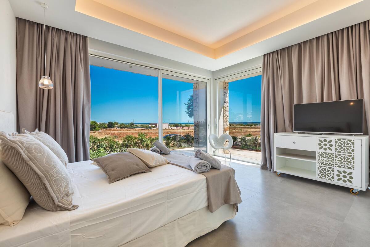 A modern bedroom with a large bed, a TV on a white cabinet, and a view of the landscape through large windows.