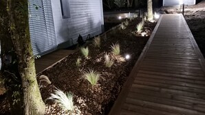 A wooden pathway leading to a white building with a garage door, surrounded by trees and illuminated by ground lights. - Very bright Scandinavian chalet located 3 min. from the lake and 10 min. of the (BISCARROSSE)