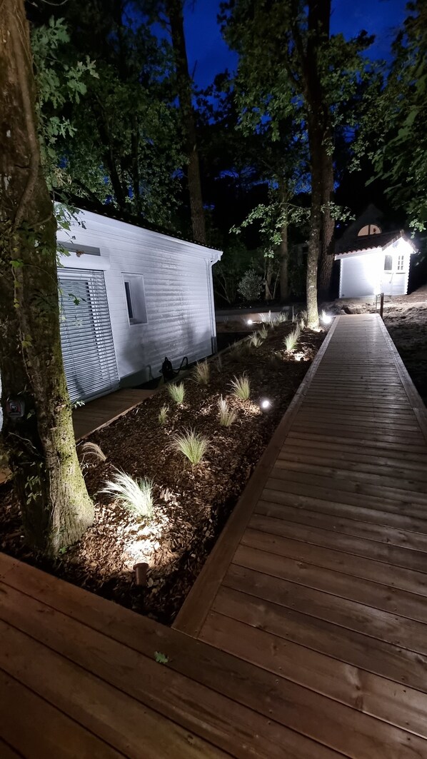 Un vialetto in legno conduce a un edificio bianco con un'autorimessa, immerso nel verde e illuminato da luci a terra.