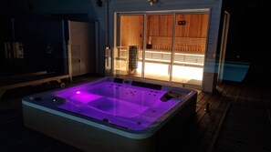 Un bain Ă remous avec une terrasse en bois et un sauna visible Ă travers une porte en verre.
