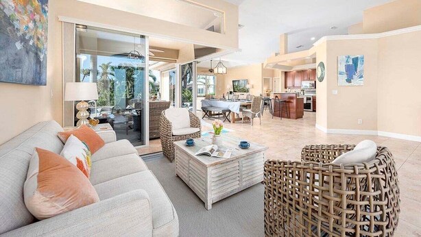 A spacious living area with a sofa, coffee table, and wicker chairs. There is a kitchen in the background with a dining table and chairs.