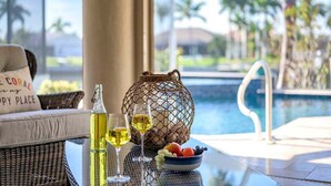 A poolside setting with a wicker chair, a glass table with a bottle and glasses, a decorative vase, and a bowl of fruit.