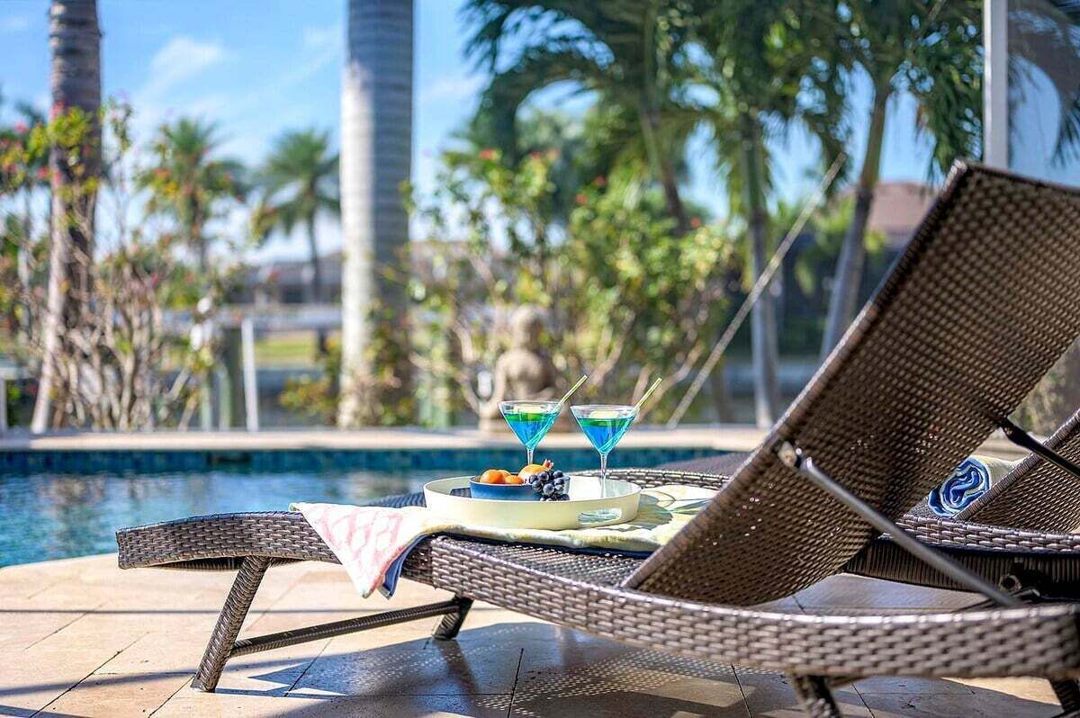 A poolside lounge chair with a tray table holding two martini glasses and a plate of fruit.