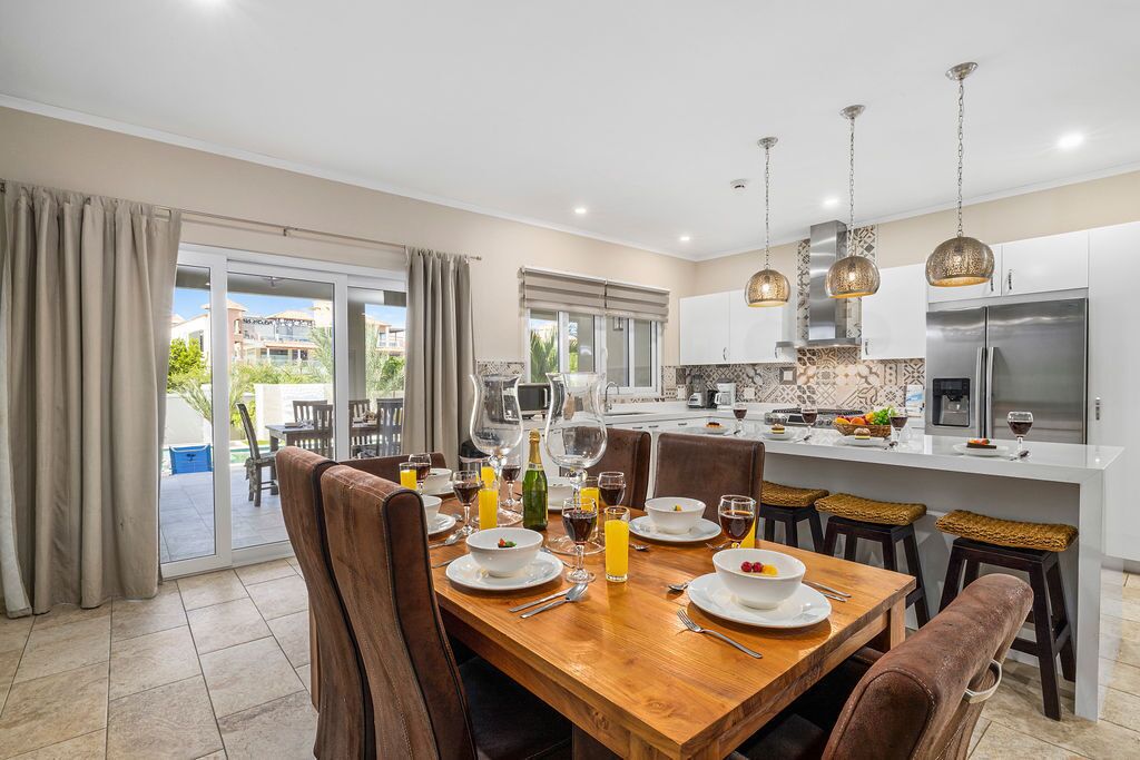Une cuisine moderne avec un coin repas, comprenant une table en bois dressée pour un repas, un grand îlot central avec un réfrigérateur et une vue sur l’extérieur.