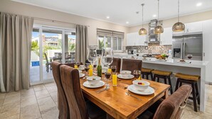 A modern kitchen with a dining area, featuring a wooden table set for a meal, a large island with a refrigerator, and a view of the outdoors.