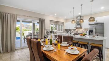 Une cuisine moderne avec un coin repas, comprenant une table en bois dressée pour un repas, un grand îlot central avec un réfrigérateur et une vue sur l’extérieur.