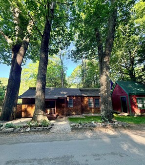 A cabin with a porch, a stone wall, and a red building in the background.