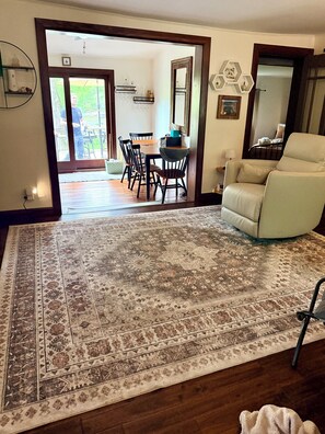 Un salón espacioso con un gran tapete estampado, una zona de comedor con sillas y una mesa, un sillón color beige, y vista hacia el comedor a través de un arco abierto.