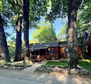 A rustic cabin with a porch, surrounded by large trees and a gravel driveway. - Charming & Cozy River Front Cabin- Close to ski mountains. Ski,Fish,Swim & Hike! (Dummerston)