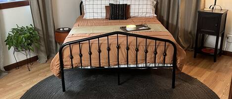 Un dormitorio con una cama centrada sobre un tapete circular, una mesita de noche con una lámpara, una planta en maceta y una ventana con cortinas.