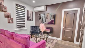 Ein Wohnzimmer mit einem rosafarbenen Sofa, einem runden Couchtisch und einem dunklen Sessel. Eine Tür mit brauner Verkleidung und ein großes Fenster mit Jalousien sind ebenfalls vorhanden.