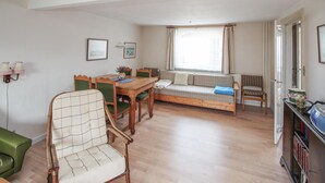 Ein gemütliches Zimmer mit einem Holztisch, grünen Sesseln, einem karierten Sofa und einem Fenster mit Vorhängen.