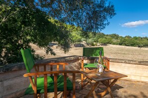 Une table et des chaises en bois sont disposées à l’extérieur, avec une bouteille et un verre sur la table, le tout entouré de verdure et sous un ciel dégagé.