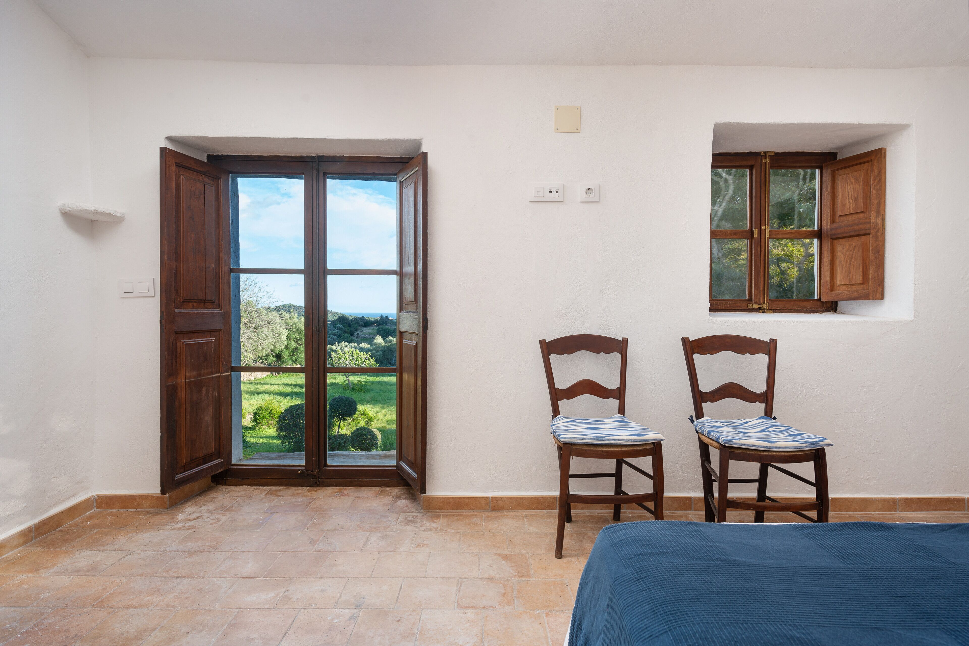 Una stanza con un'ampia porta a vetri scorrevoli che dà su un balcone, due sedie di legno con cuscini decorati e una piccola finestra con persiane di legno.