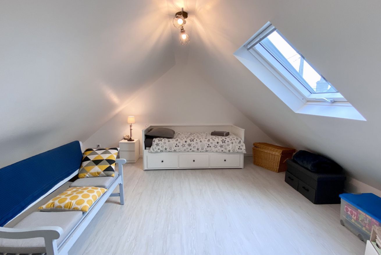 Ein kleines, gut beleuchtetes Schlafzimmer im Dachgeschoss mit schrägen Wänden, einem Dachfenster, einem Bett mit gemustertem Überwurf, einem blauen Sofa und einem Nachttisch mit Lampe.
