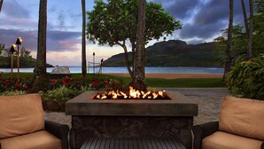 A stone fire pit with a table in the center, surrounded by wicker chairs with cushions.