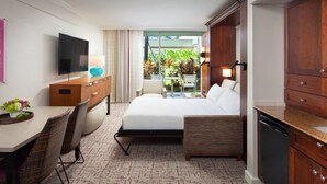 A hotel room with a bed, a desk, a television, a dining area, and a view of greenery outside.