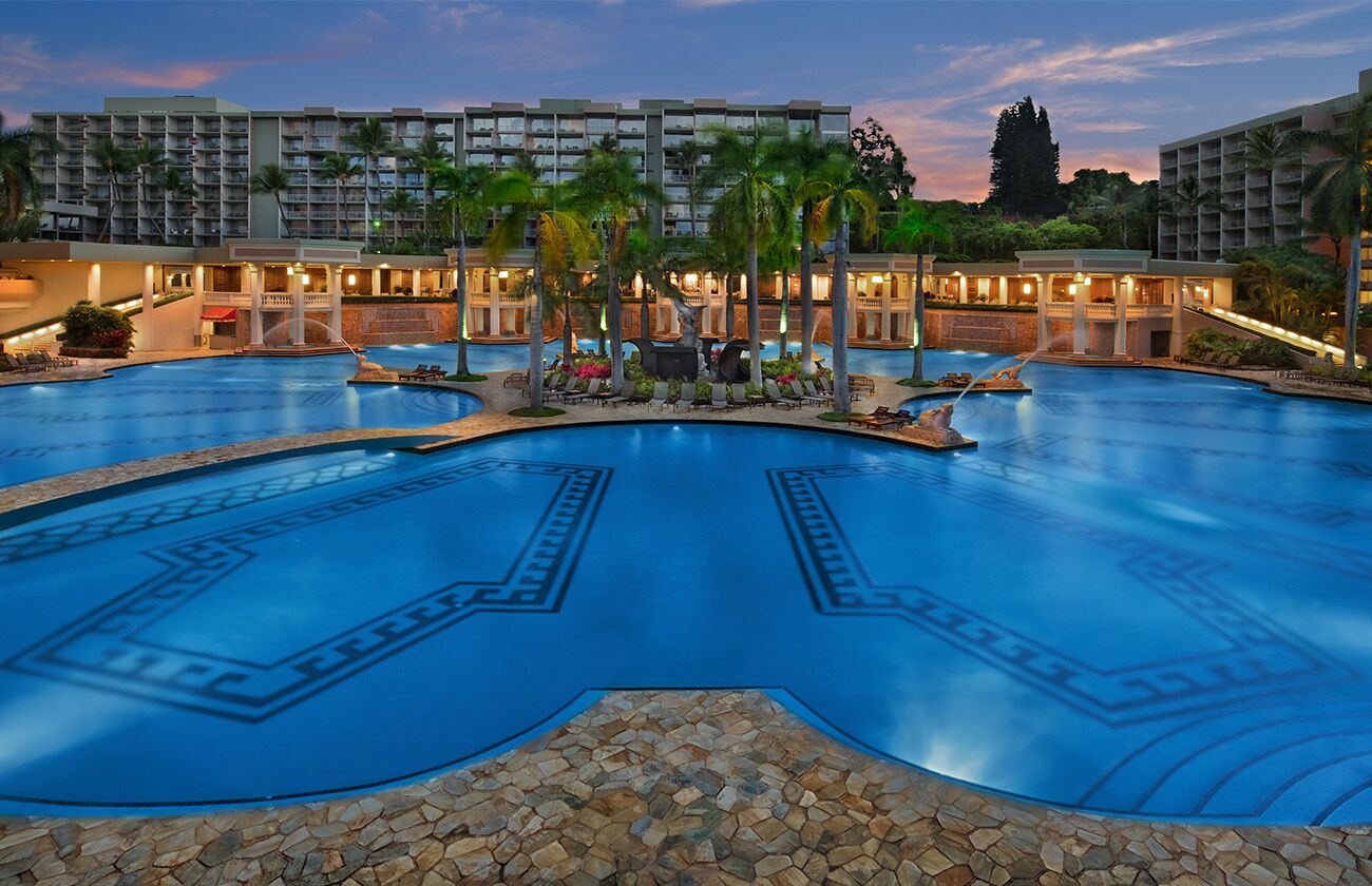 Una gran piscina circular con un diseño geométrico en el suelo, rodeada de palmeras y edificios de hotel al fondo.
