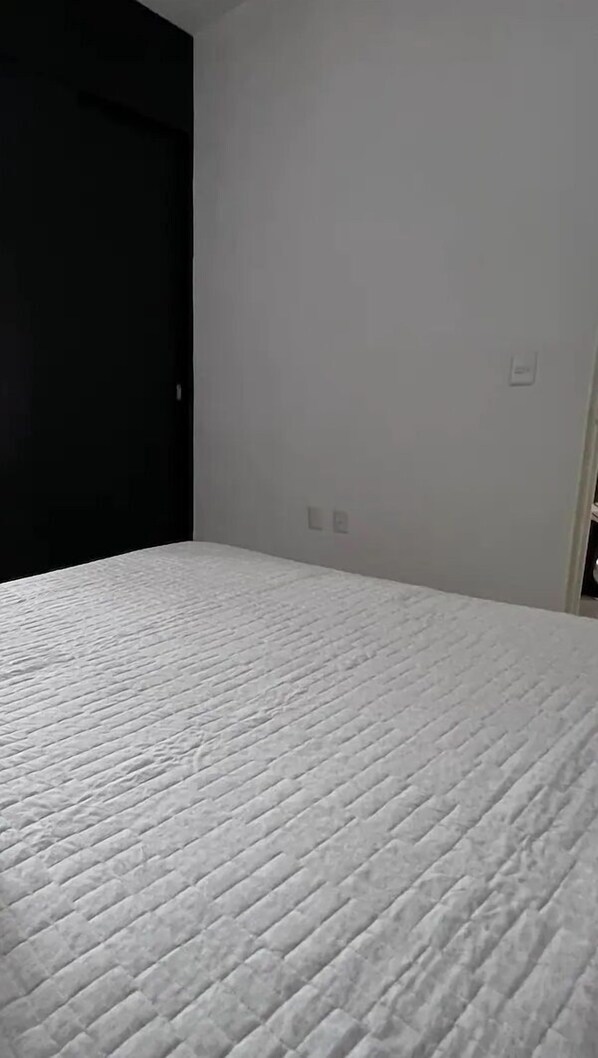 Habitació amb un llit, una porta fosca i una paret de color clar amb dos interruptors.