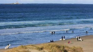 Una playa con surfistas y un faro a lo lejos.