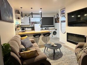 Una sala de estar moderna con sofá, mesa de centro y televisor. Cuenta con una zona de cocina con refrigerador, microondas y lavaplatos.