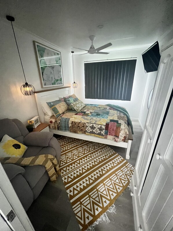 Ein Schlafzimmer mit einem Bett, einem grauen Sofa, einem gemusterten Teppich, einem Deckenventilator und einem Fenster mit Jalousien.
