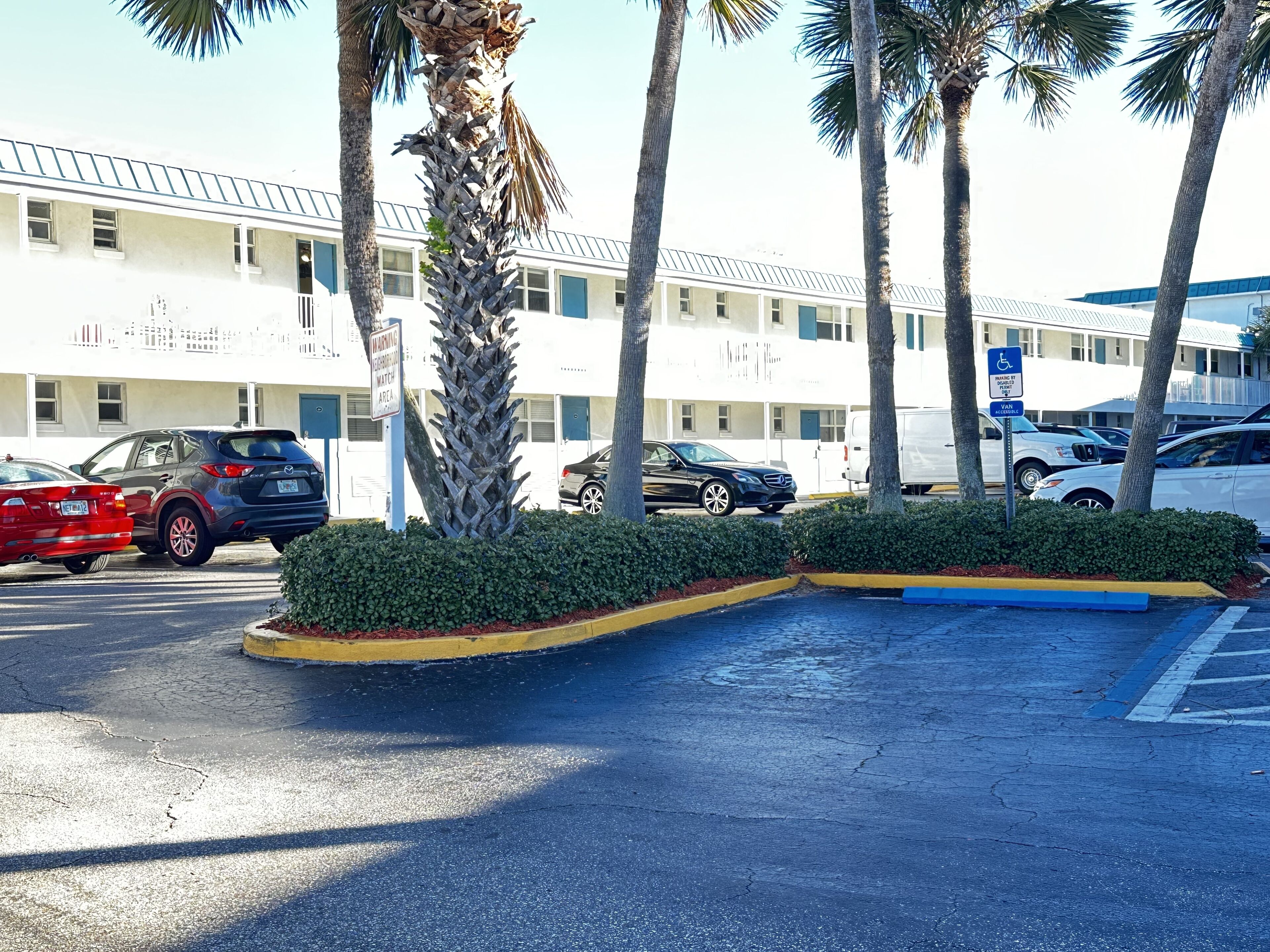 A parking lot with several cars, palm trees, and a building with multiple windows and a flat roof.
