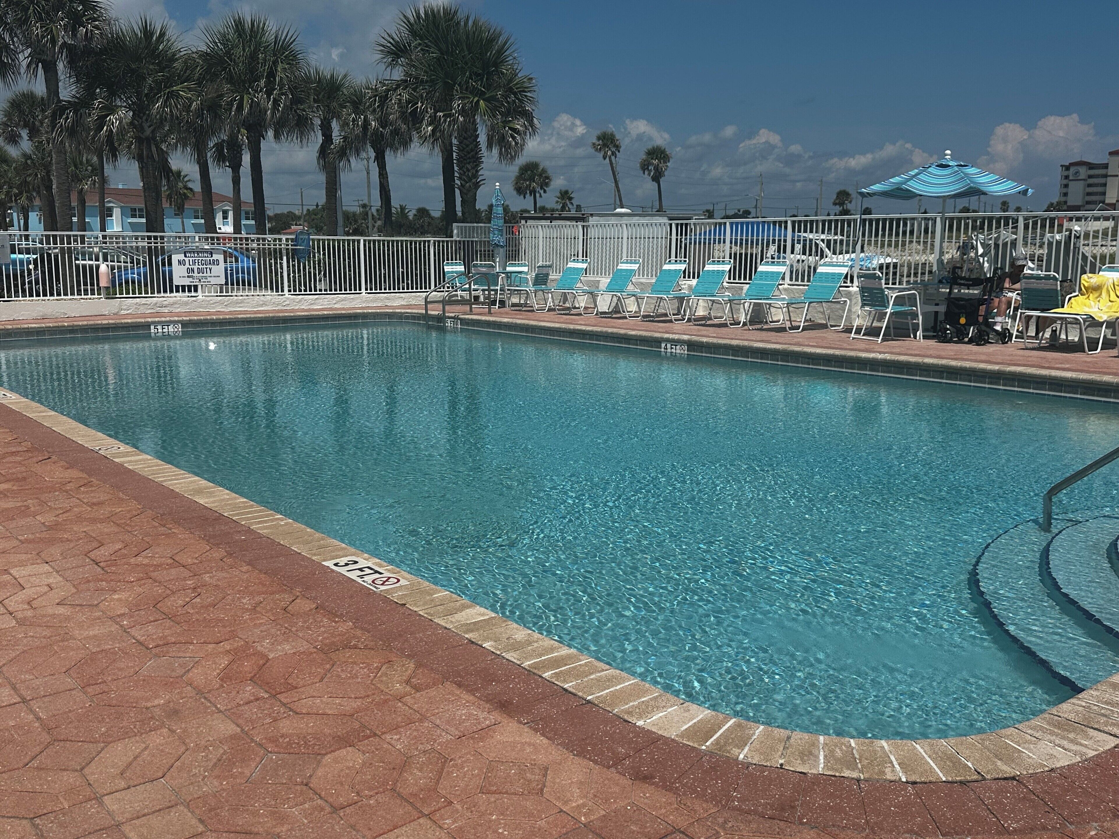 A swimming pool with lounge chairs and umbrellas, surrounded by palm trees and a fence.