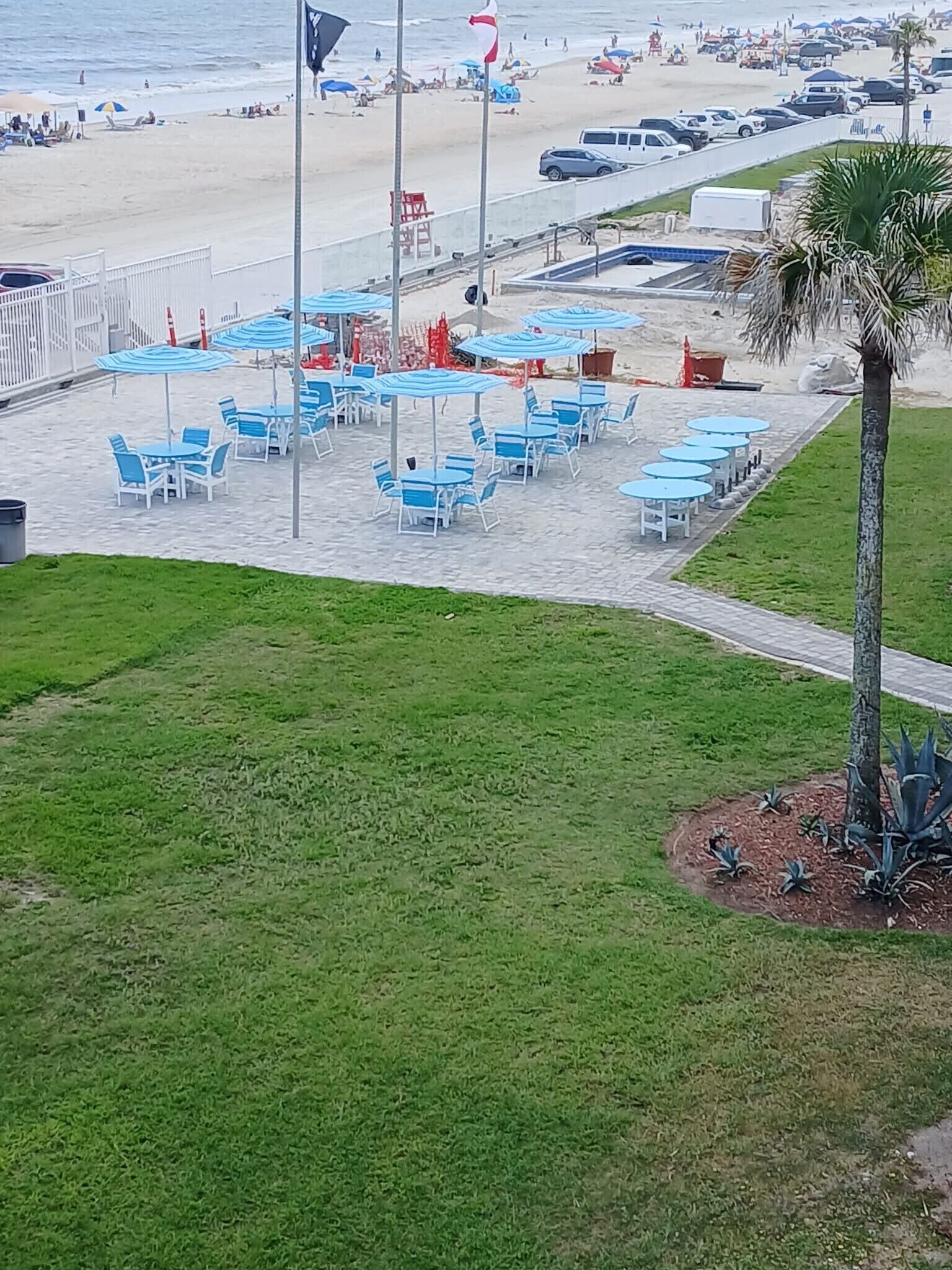 En uteplats vid stranden med blå och vita bord och stolar, ett palmträd och en flaggstång.