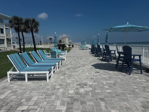 A beachside promenade with lounge chairs and umbrellas, a paved walkway, and a view of the ocean.