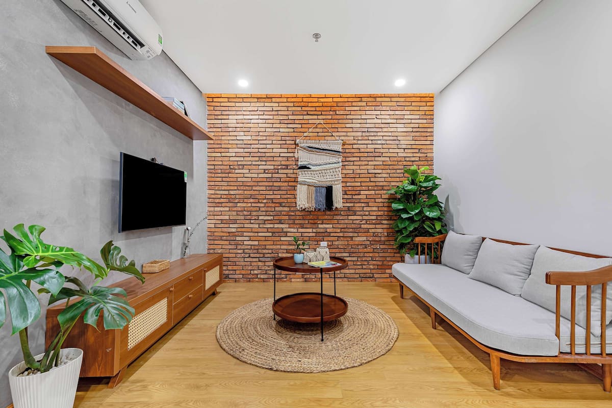 A modern living room with a brick wall, a flat-screen TV, a wooden sofa, a wooden console table, and a potted plant.