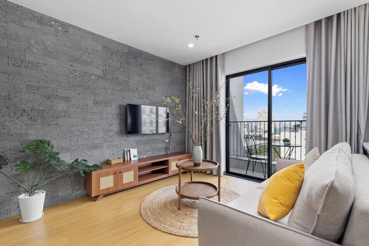 A modern living room with a grey concrete wall, wooden furniture, a flat-screen TV, a large window with a city view, and a balcony.