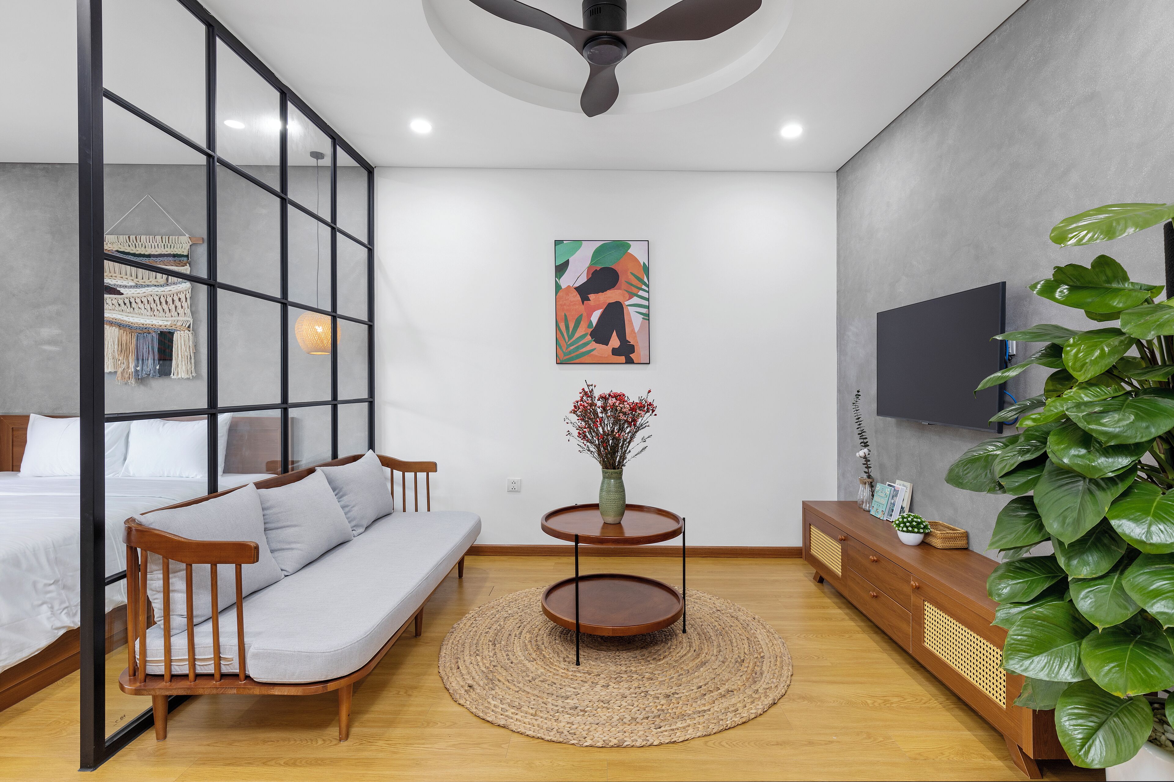 Un dormitorio moderno con un sofá de madera, una pequeña mesa redonda con un jarrón, una planta verde grande y un televisor montado en la pared.