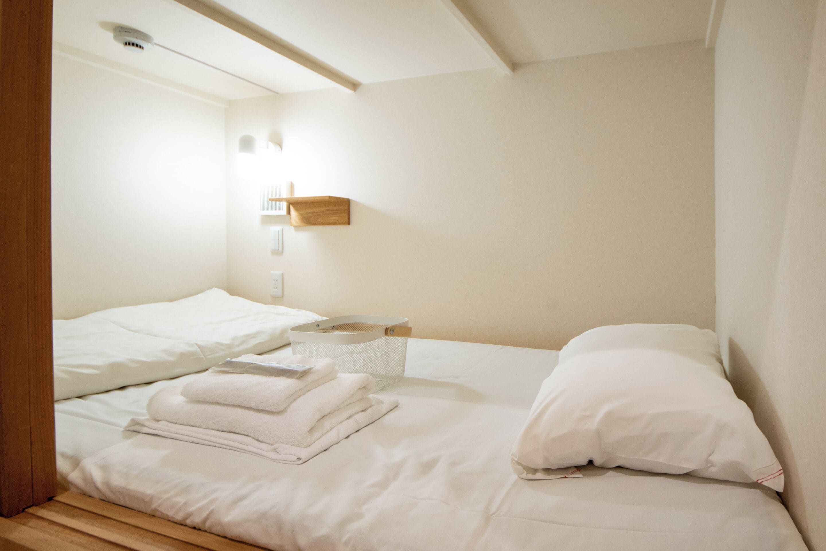 A neatly made bed with white linens and a folded towel, a bedside table with a basket, and a wall-mounted light fixture.