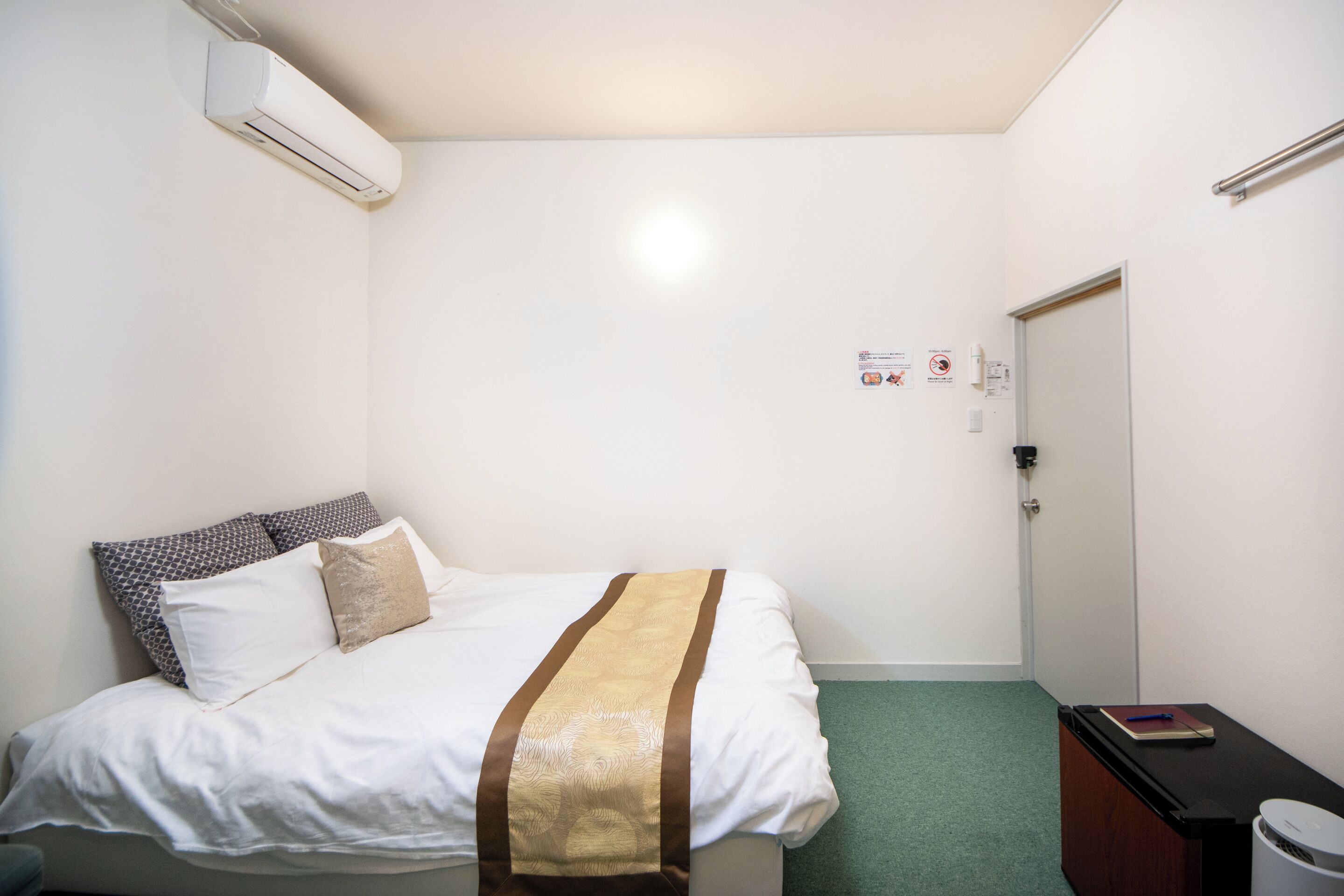 A hotel room with a bed, a desk, a trash bin, and an air conditioning unit.