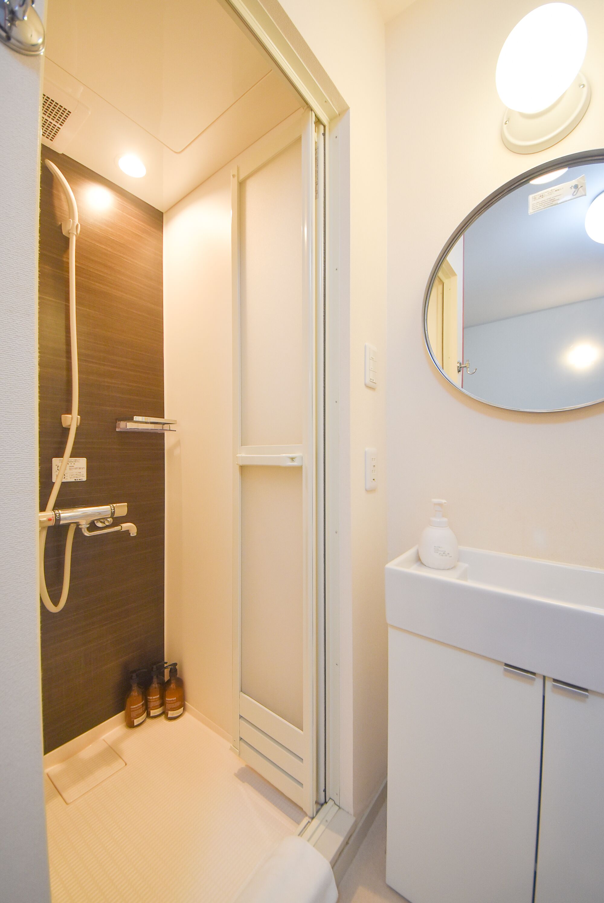Un baño con cabina de ducha de vidrio, un lavamanos blanco con un espejo encima y una botella de champú en la encimera.