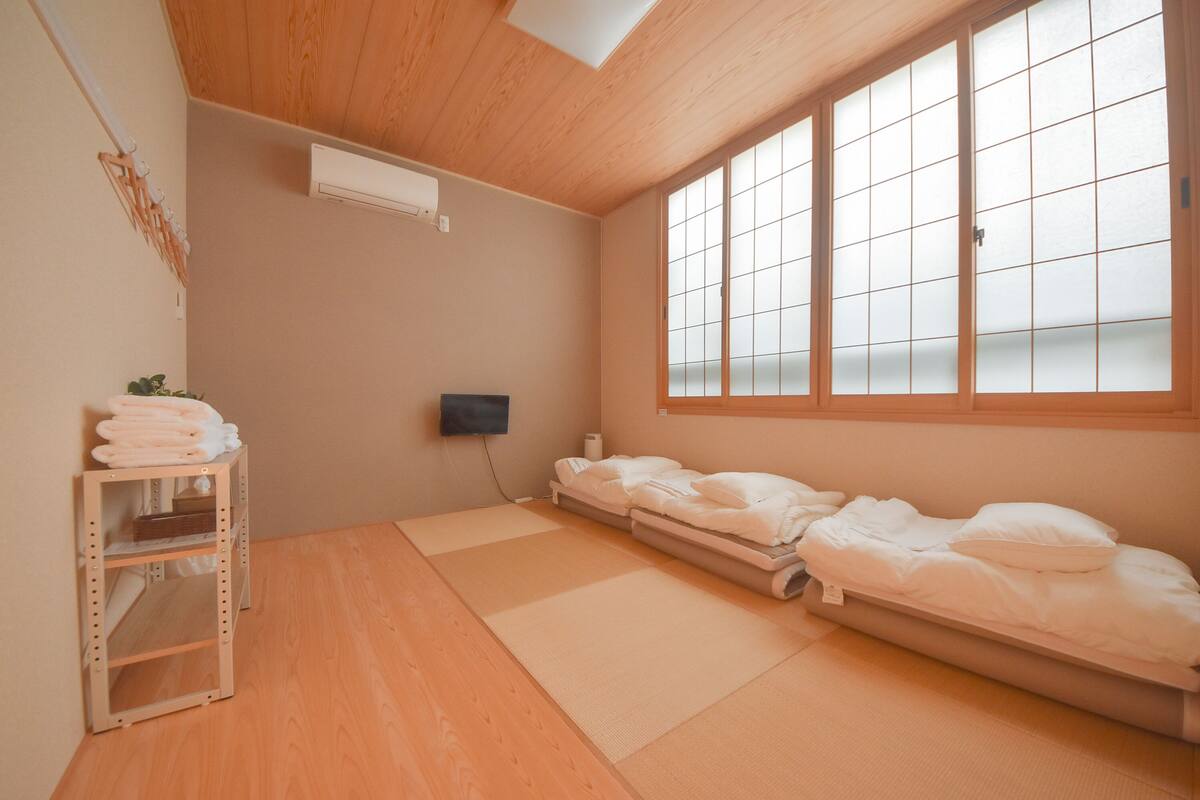 A room with a wooden ceiling, a large window with frosted glass, a bed with white bedding, a shelf with towels, and a wall-mounted air conditioner.