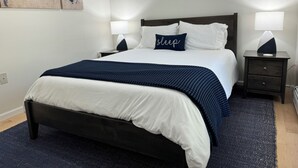 A neatly made bed with a dark blue bedspread and pillows, two bedside lamps, a world map on the wall, and a dark wooden nightstand with drawers. - Charming home near PSU - built for memorable game days and family visits (State College)