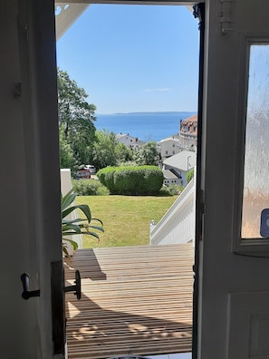 Une vue depuis la porte d’une pièce, donnant sur un balcon qui surplombe une zone herbeuse, une haie et une étendue d’eau au loin.