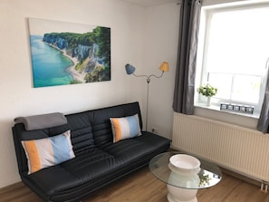 Un salon avec un canapé noir, une table basse en verre, un tableau représentant un paysage côtier, une lampe sur pied et une fenêtre avec des rideaux.
