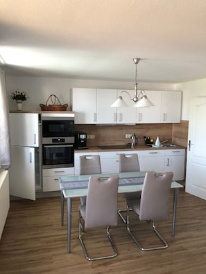 Una cocina moderna con armarios blancos, electrodomésticos de acero inoxidable, una mesa de vidrio con sillas y una lámpara colgante.