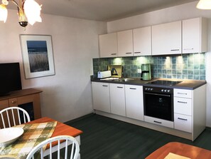 Kompakti keittiö, jossa on valkoiset kaapit, musta uuni, kahvinkeitin sekä ruokailutila, jossa on pöytä ja tuolit.