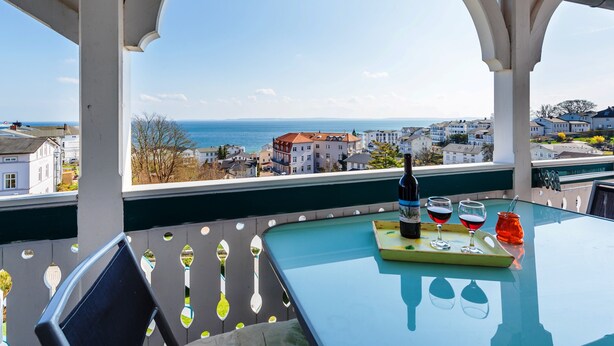 En balkon med et bord, en flaske og glas, med udsigt over en kystby.