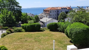 A residential area with a prominent building featuring a red roof and multiple windows, surrounded by lush greenery and a clear view of the sea.