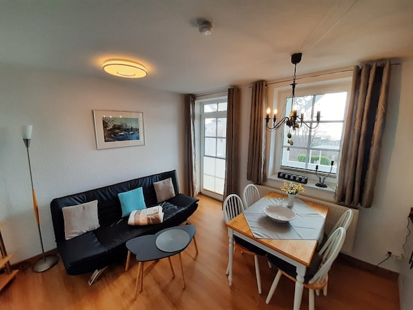 A cozy living area with a black sofa, a small round table, and a dining table with chairs. A chandelier hangs above the dining area, and a window with curtains is visible.