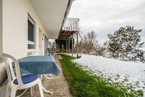 Een overdekt terras met een tafel en stoelen, een houten pergola, en een besneeerd landschap met bomen.