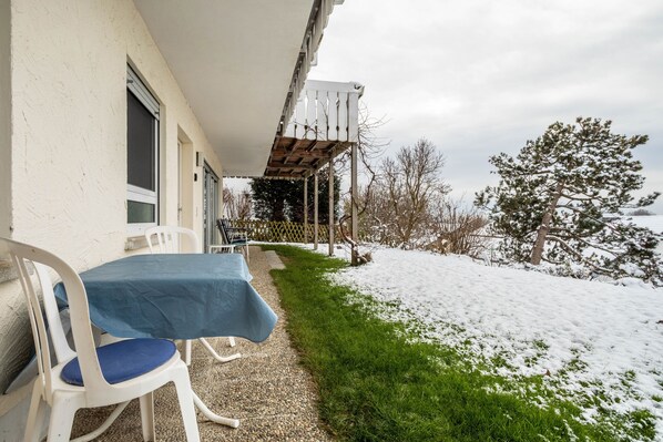 A covered outdoor seating area with a table and chairs, a wooden pergola, and a snow-covered landscape with trees.