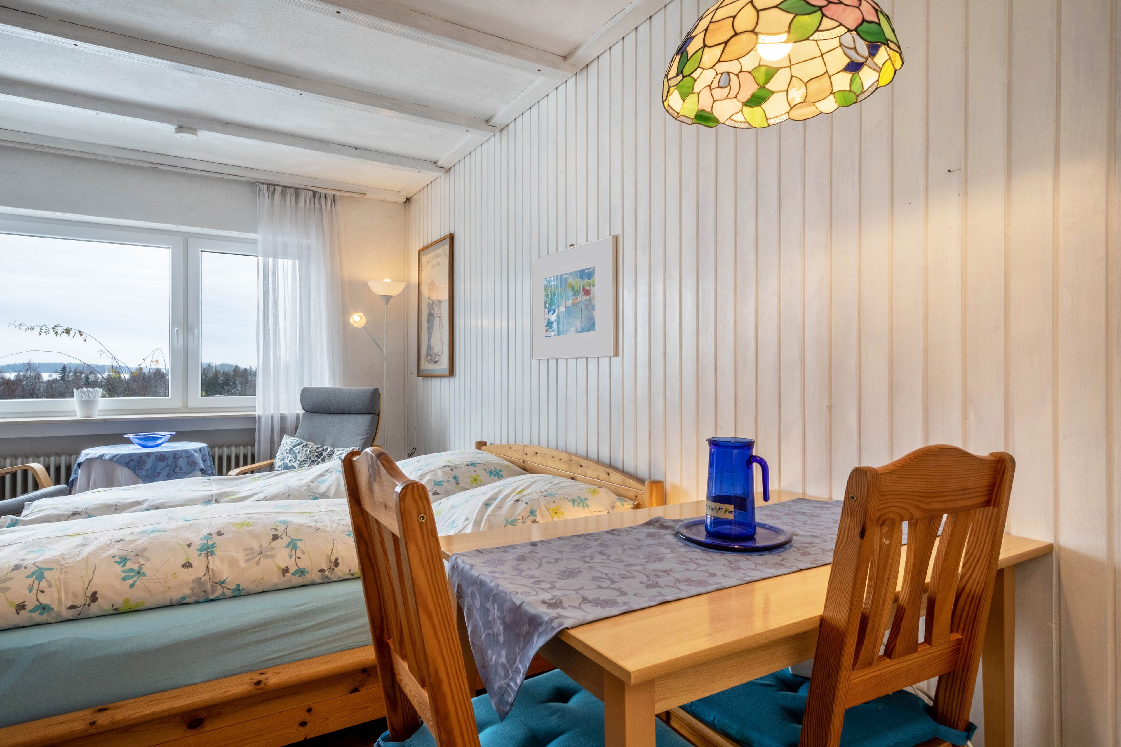 Ein Zimmer mit zwei Betten, einem Tisch mit Stühlen, einem Buntglasfenster und Blick ins Freie.