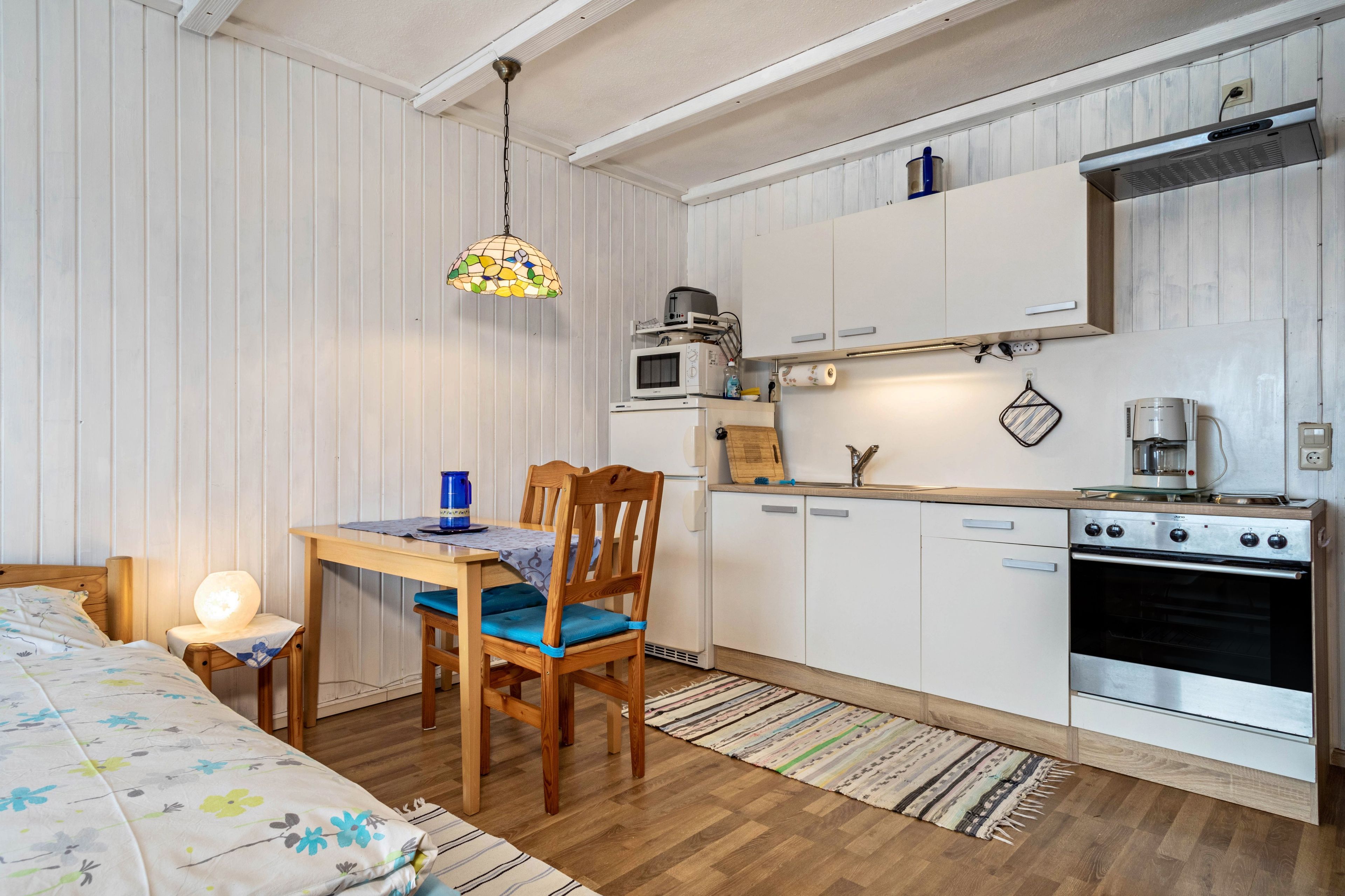 Cocina compacta con alacenas blancas, una mesa de comedor de madera, una silla, una lámpara colgante y un horno empotrado.