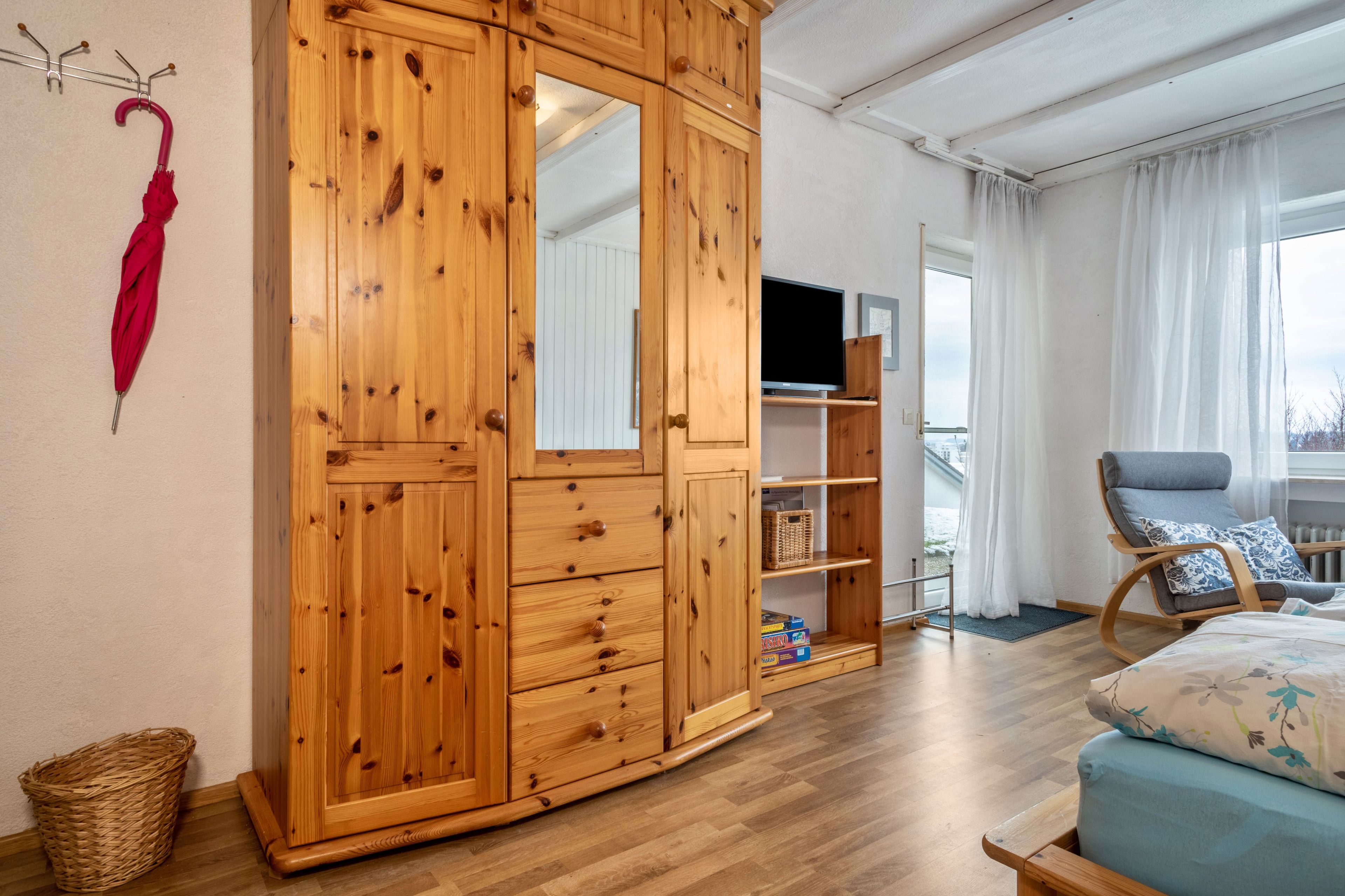 Een houten garderobe met meerdere lades, een televisie op een standaard, een rieten mand en een blauwe bank met bloemenkussens.